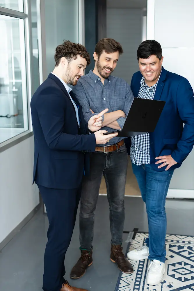 Three colleagues standing together in an office hallway smiling and looking at a laptop.
