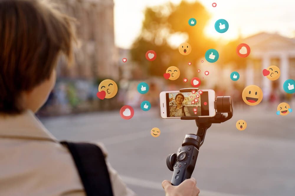 influencer marketing_Teen girl blogger vlogger record vlog streaming video hold phone on selfie stick in urban city. Young female vlogger shoot social media blog on smartphone get likes emoji, over shoulder closeup view.