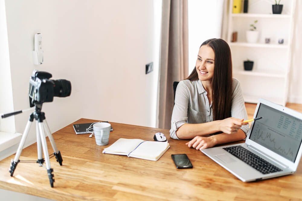 influencer marketing_Portrait of a woman with a laptop. She points to the laptop screen, looks at the camera on a tripod. Recording video lessons, video blogs