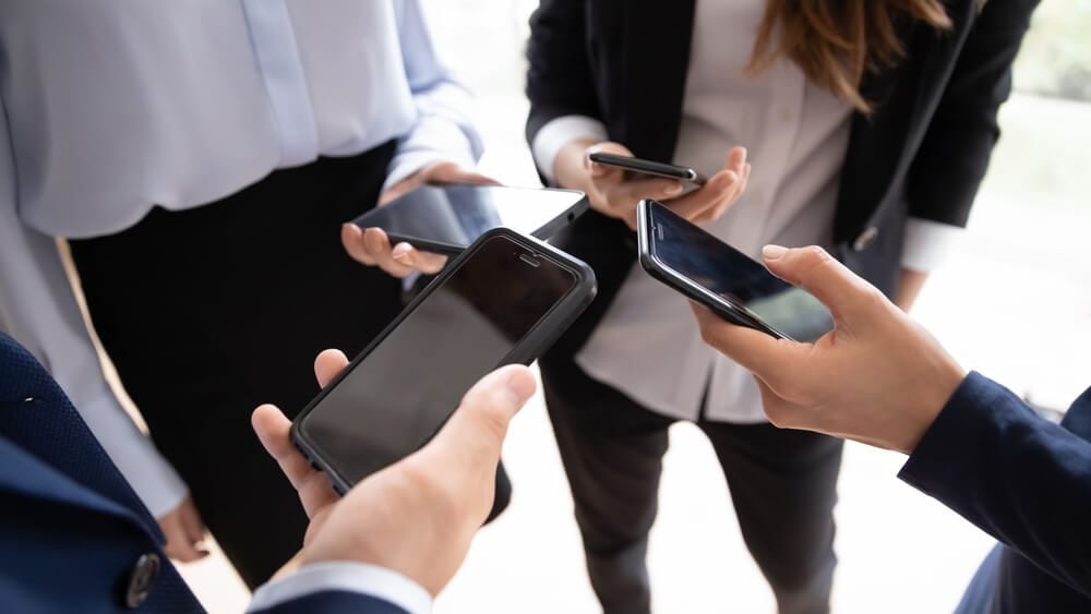 mobile app_Four entrepreneurs standing in circle close up view on hands holding smart phones. Solving business issues distantly, corporate chat app, business application usage, phubbing, devices overuse concept