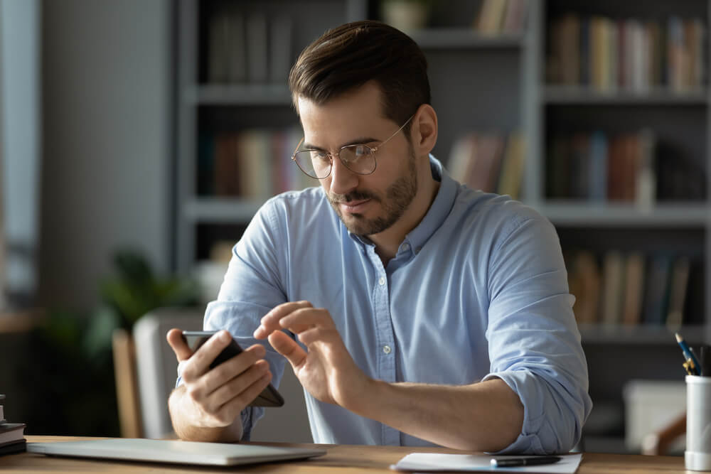 solving issues mobile app_Serious businessman wearing spectacles using mobile phone sit at desk in cozy office. Manager solve issues with client remotely by modern wireless device, surfing web, learn new business app concept