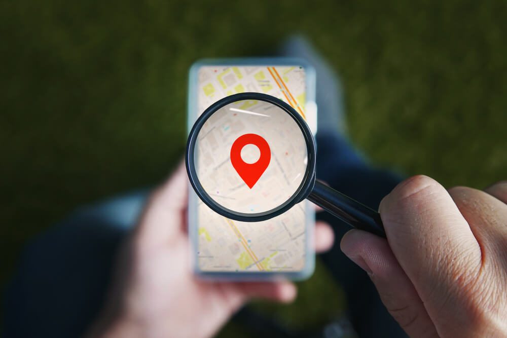 local search_Shot of male hands holding magnifying glass with the red icon of geo-location and looking through it to the smartphone with online city map on the screen. Concept of using mobile net for navigation