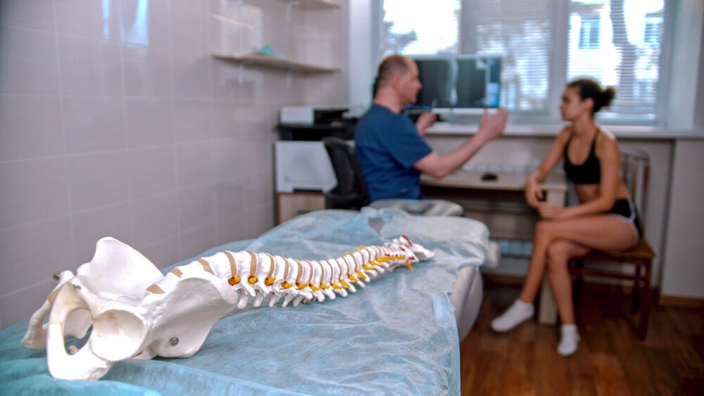 chiropractic_Chiropractic treatment - the doctor inspecting the young woman before the session - model of the human spine placed on the couch in the cabinet