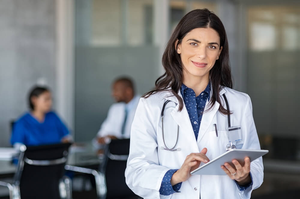 doctors_Portrait of beautiful mature woman doctor holding digital tablet and looking at camera. Confident female doctor using digital tablet with colleague talking in background at hospital. Latin nurse.