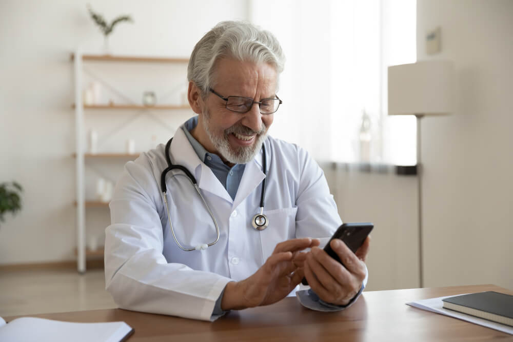 physician_Happy old doctor holding smart phone texting message video calling for online telemedicine consultation in medical office. Smiling senior male physician using mobile healthcare app on cell technology.
