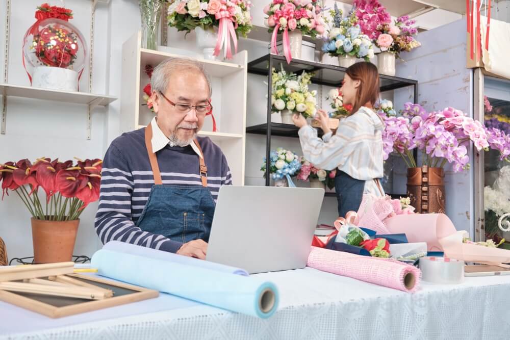 florists_Asian old male florist owner working with a laptop on website for online business, a young beautiful female shopkeeper arranging fresh blossom behind, colorful flower shop store, e-commerce SME.