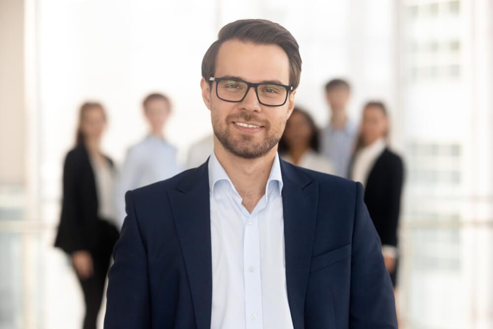 fractional marketing director_Confident business man coach in suit director company owner professional manager looking at camera with team on background, smiling male ceo leader banker employer posing in office, headshot portrait