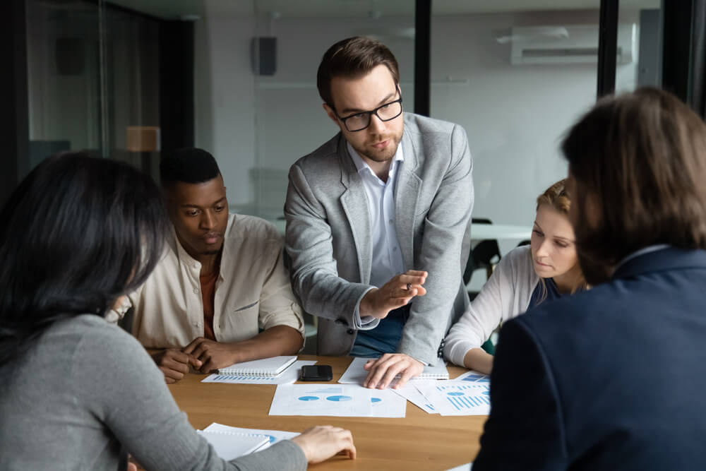 marketing officer_Focused confident young employee in eyeglasses giving arguments about marketing strategy or sharing own business ideas with focused motivated mixed race workgroup at brainstorming meeting in office.