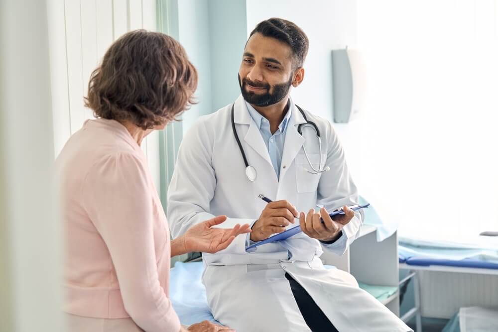 doctors patient_Indian male doctor consulting senior old patient filling form at consultation. Professional physician wearing white coat talking to mature woman signing medical paper at appointment visit in clinic.