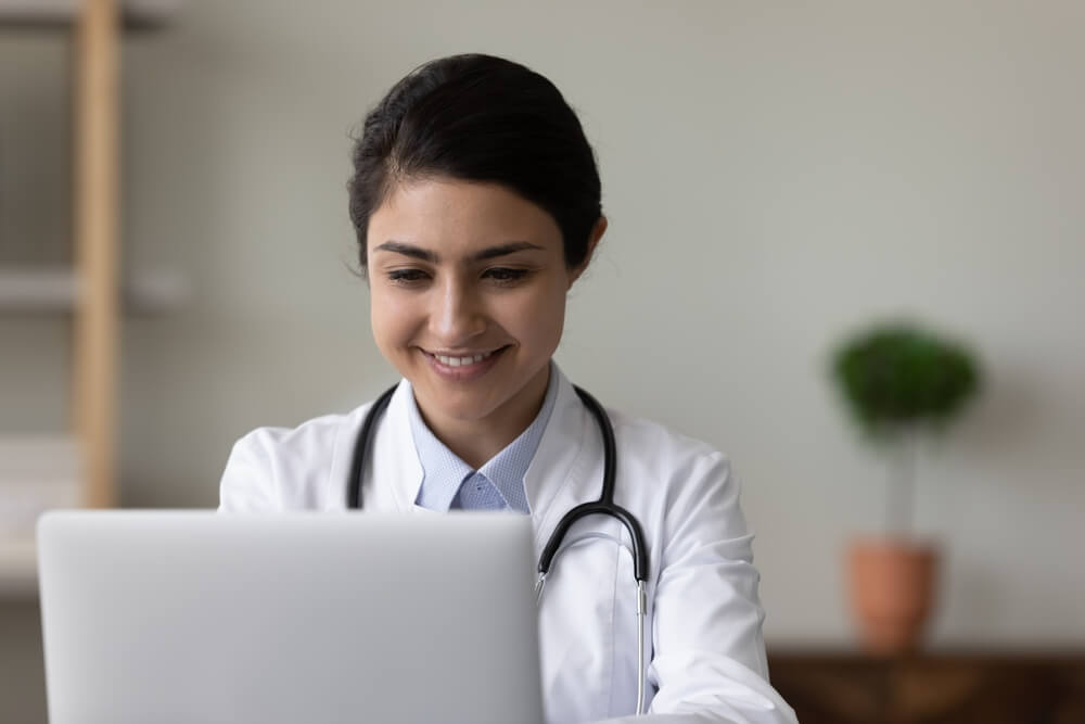 healthcare email_Happy female Indian doctor chatting online with patient, giving virtual consultation, making video call on laptop computer. Young therapist working at computer in clinic office, smiling at screen