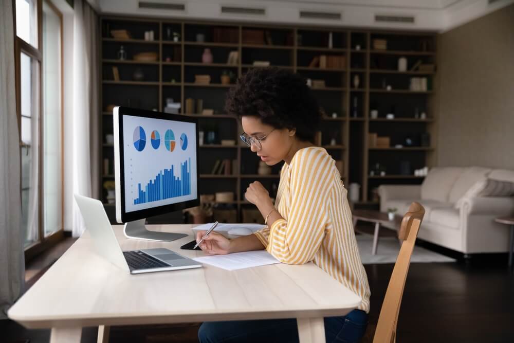 Market Analytics_Concentrated young African American businesswoman employee worker in eyeglasses analyzing online sales statistics data on computer, reviewing marketing research, working with documents at home office.