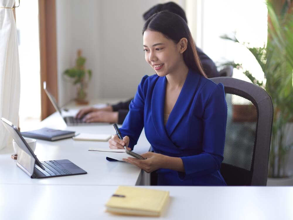 CMO_Happy Asian female businesswoman looking at the tablet screen, taking note on desk at office workplace.