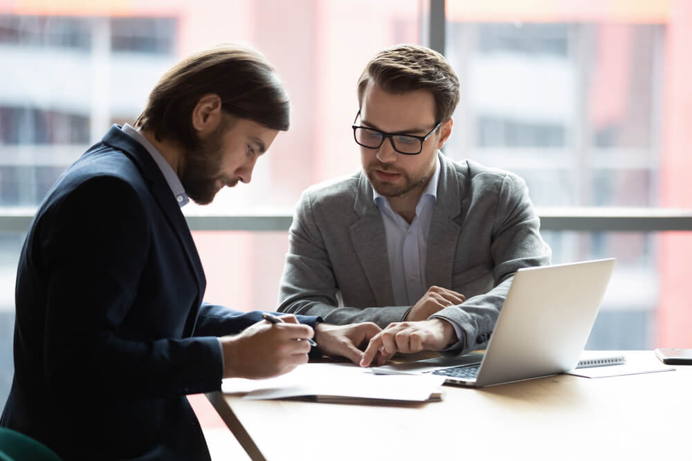 marketing consultant_Focused young businessman signing agreement with skilled lawyer in eyeglasses. Concentrated financial advisor showing place for signature on paper contract document to male client at meeting in office