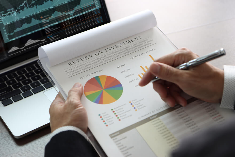 ROI_Closeup on businessman or analyst hands holding financial statements reviewing on return on investment, ROI, investment risk analysis in front of laptop computer with graphs and related information