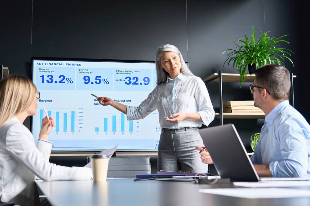 revenue team_Mid age senior Asian business woman ceo executive manager showing to colleagues team income revenue data figures planning on big screen in modern office on business seminar workshop.