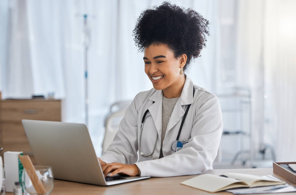 healthcare email_Black woman, doctor and laptop with smile for healthcare, email or telemedicine by work desk at the hospital. African American female medical expert smiling on computer in medicare research at clinic