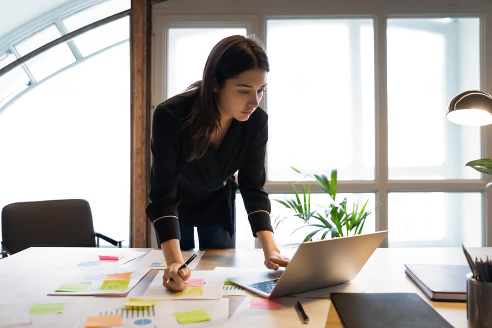 demand_Confident millennial indian female business analyst financial advisor preparing statistic report studying documents on work desk, browsing information online using pc, writing out notes on paper sheet