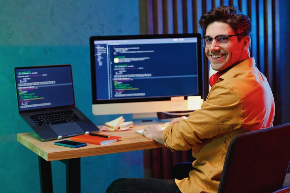 web development_Side back view smiling young full-stack developer software engineer IT specialist programmer man in shirt work at home writing code on laptop pc computer checking database. Program development concept
