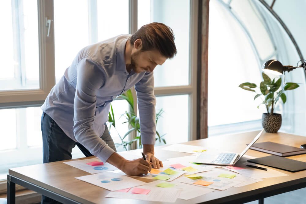 demand generation_Thoughtful serious young businessman employee standing at desk focused on paperwork, making changes correcting mistakes in marketing plan by hand, analyzing client customer demands using infographics