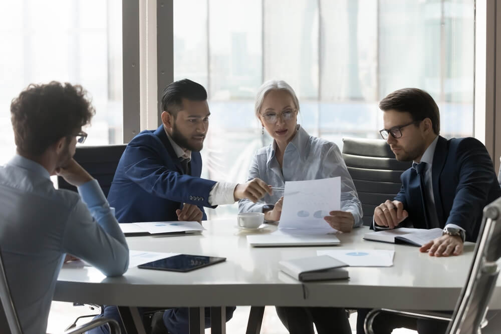 SEO_Serious multiracial older and young businesspeople gathered in boardroom discuss financial statistics, analyze sales report, forecasting work together at office meeting. Teamwork, negotiations concept