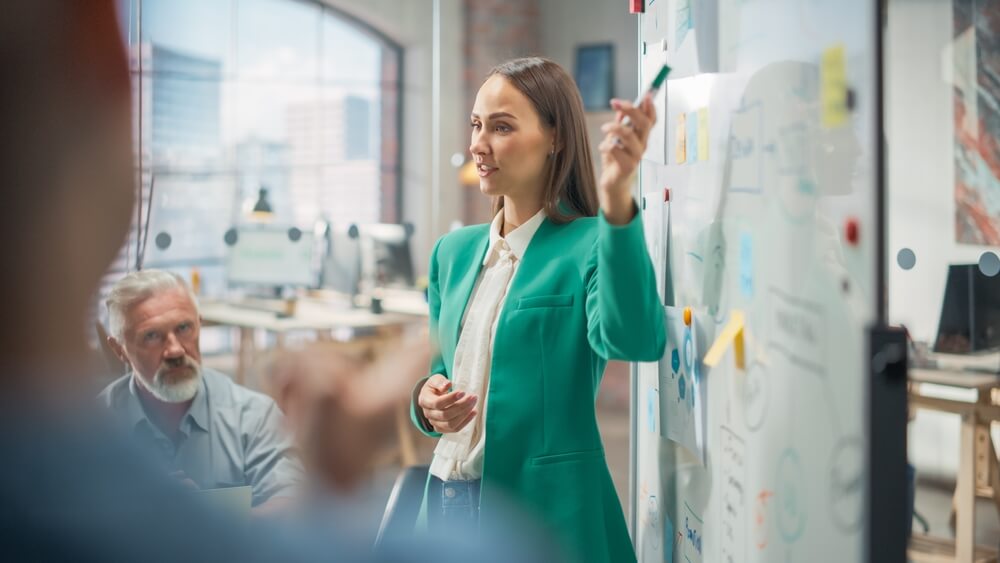 FCMO_Female CEO Using Whiteboard in Office to Answer Questions and Give Feedback to her Team During a Meeting. Portrait of Beautiful Woman Doing Presentation in Creative Agency