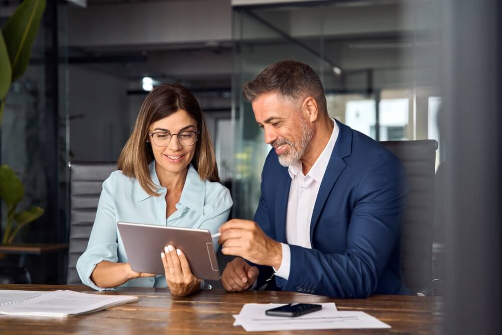 SEO consultant_Team of diverse partners mature Latin business man and European business woman discussing project on tablet sitting at table in office. Two colleagues of professional business people working together.