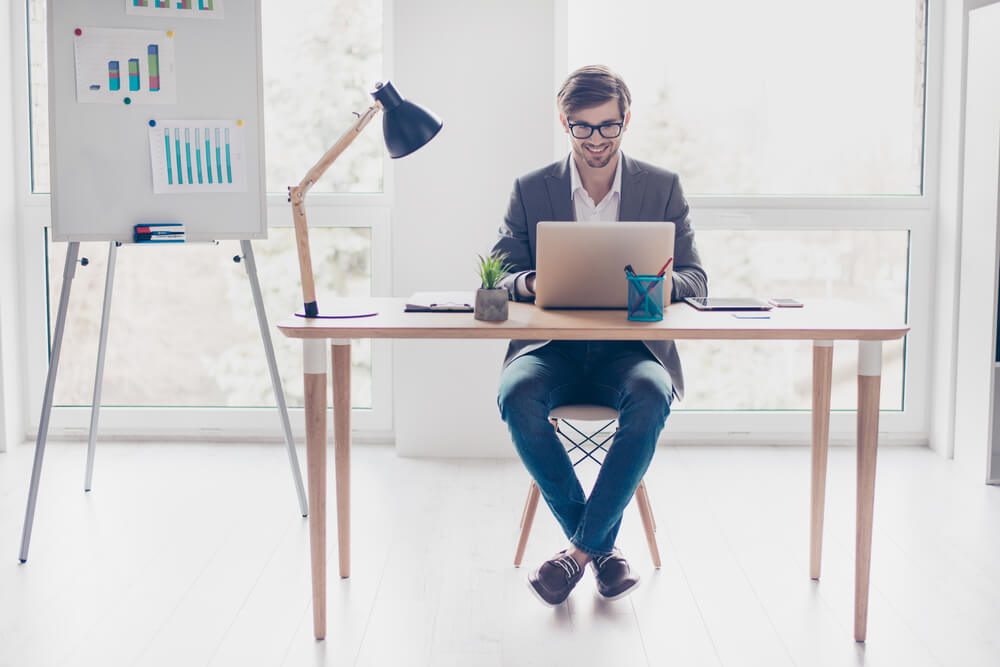 content intelligence_Portrait young handsome businessman with glasses is sitting at desk in workstation, smiling and typing on laptop.