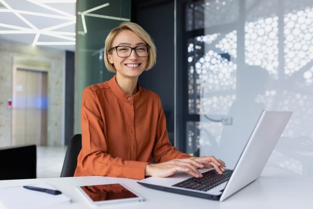 CMO_Portrait of happy and successful female programmer inside office at workplace, worker smiling and looking at camera with laptop blonde businesswoman is satisfied with results of achievements at work