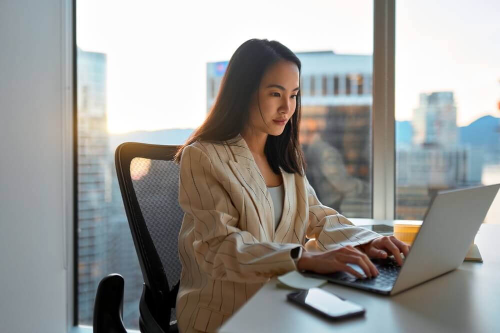 fractional CMO_Busy Asian business woman using laptop in company office. Young female digital finance professional worker using computer doing corporate analysis online management sitting at desk, city window view.