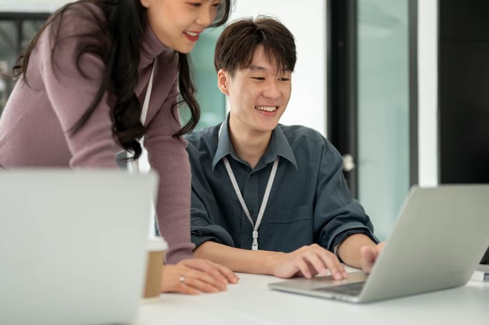 SEO_Close-up image of a professional Asian female marketing assistant is reading details on a laptop and helping her male colleague with a marketing campaign, working in the office together.