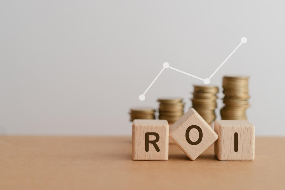 ROI_Cube block wood letters ROI and blurred money coin stack with white increasing graph including copy space. For Return on Investment financial concept