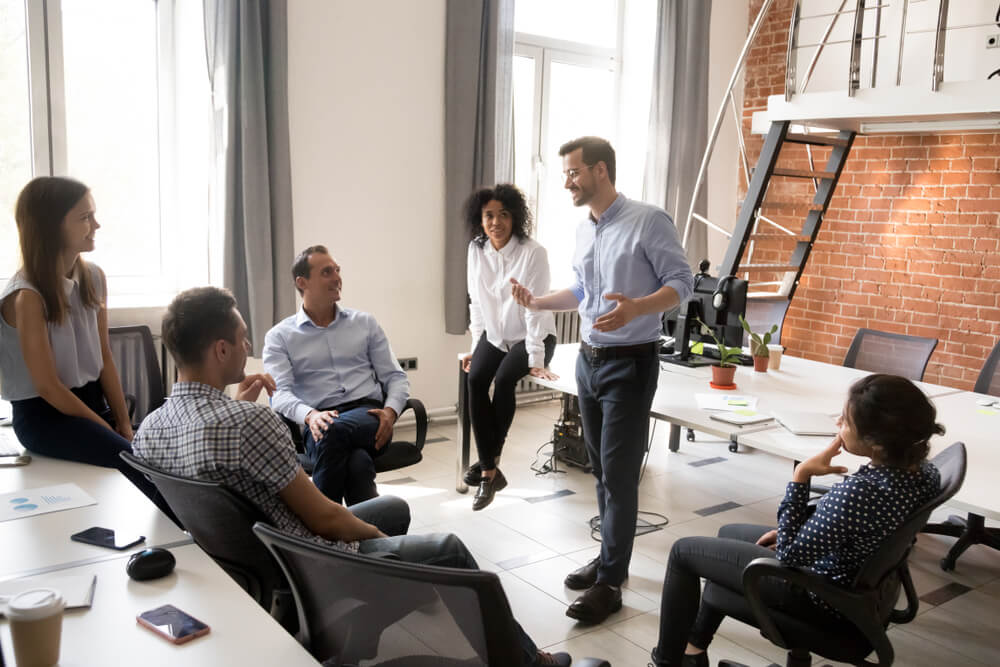 FCMO_Confident male leader, coach talking with multiracial group of office workers, having good conversation with subordinate, brainstorming, discussing business strategy, ideas, team building activity