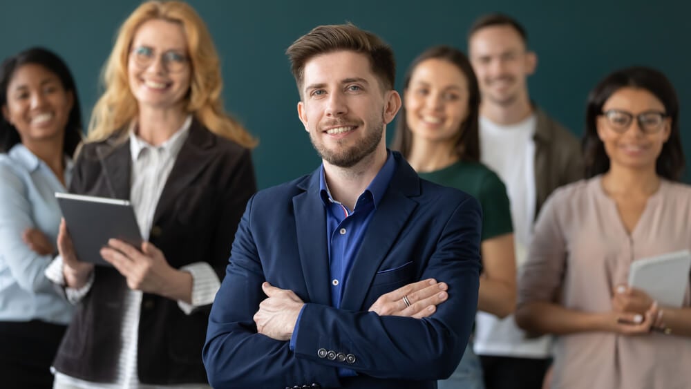 CMO_Happy confident male business leader posing with team in blurred background. Young businessman, director, manager with group of office employees looking and smiling at camera. Head shot portrait