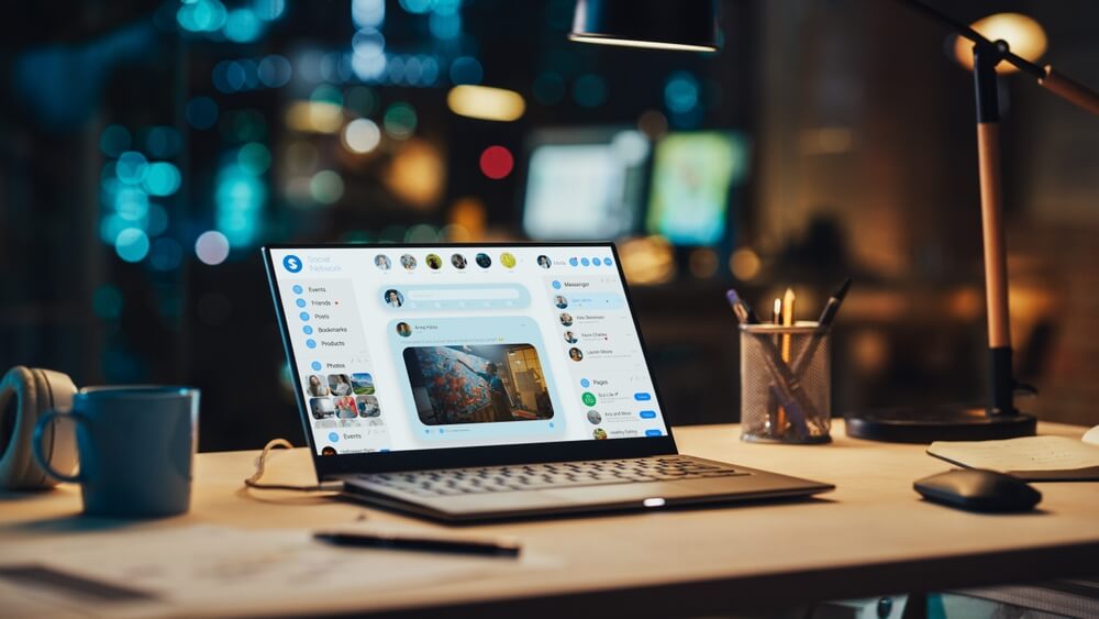 eCommerce Social Media_Laptop Standing on a Desk with a Social Media Platform Displayed on Screen. Table with Computer, Coffee Mug, Headphones and Notebook in Creative Office at Night.