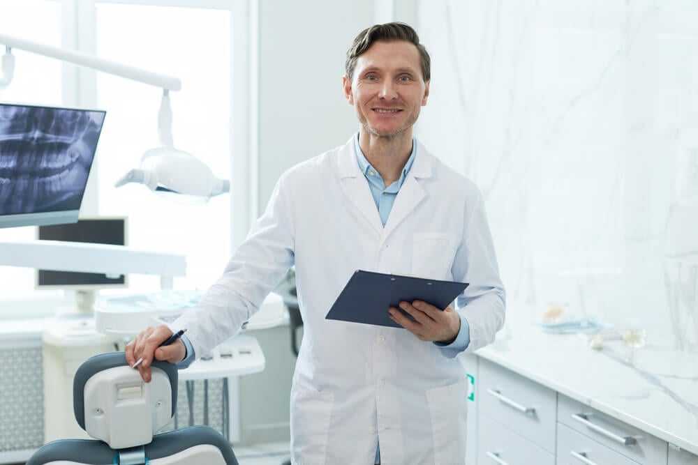 dentist_Waist up portrait of smiling male dentist looking at camera in dental clinic and holding clipboard, copy space