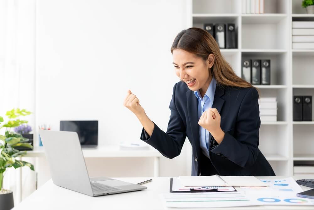 B2B success_Young Asian business woman wearing suit and using laptop on gray baclground Cheerful young woman celebrating her achievement while reading good news on laptop from home office