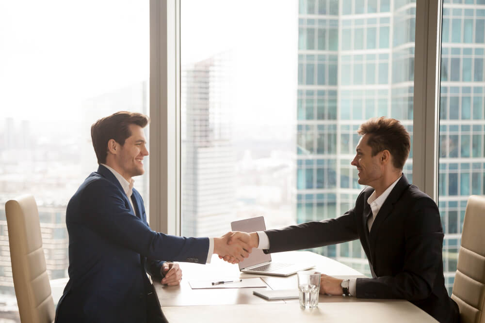 brand trust_Two happy satisfied businessmen shaking hands over desk after successful negotiations, closing sealing deal, big window city building at background, smiling partners binding agreement with handshake