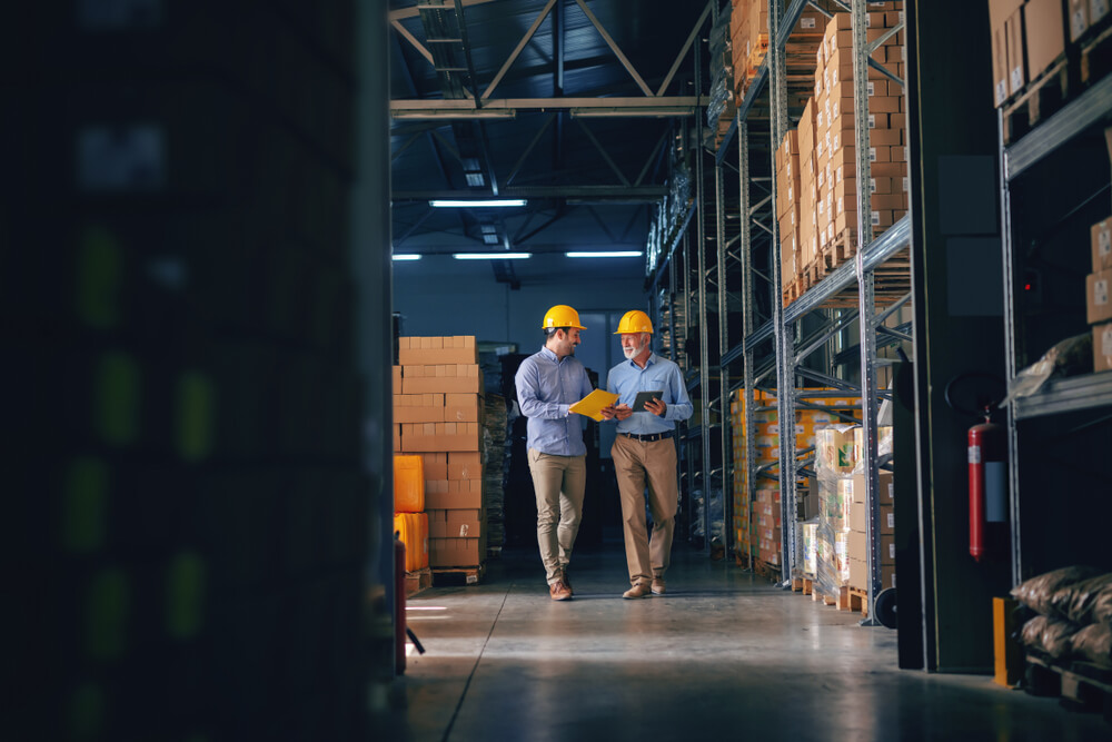 manufacturing_Two business partners in formal wear and with protective yellow helmets on heads walking and talking about business. Younger one holding folder with data while older one using tablet.