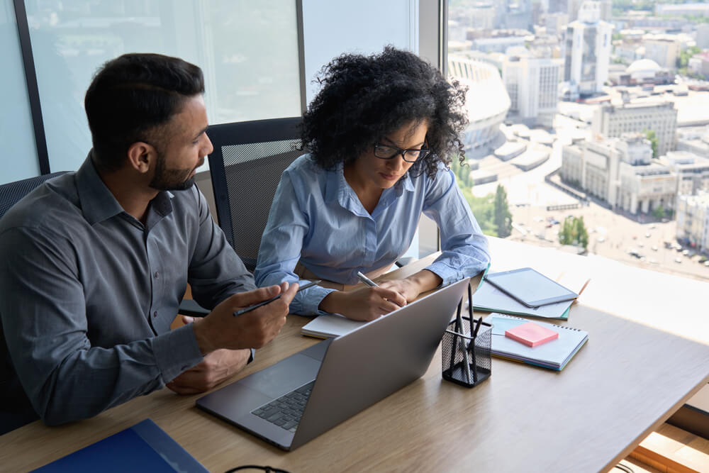 digital consulting_Multiethnic coworkers male indian mentor and female African American intern sitting at desk with laptop working together discussing project writing notes. Corporate business collaboration concept.