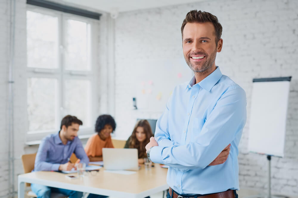 demand strategy_Happy businessman standing in the office with coworkers in the background working by the desk