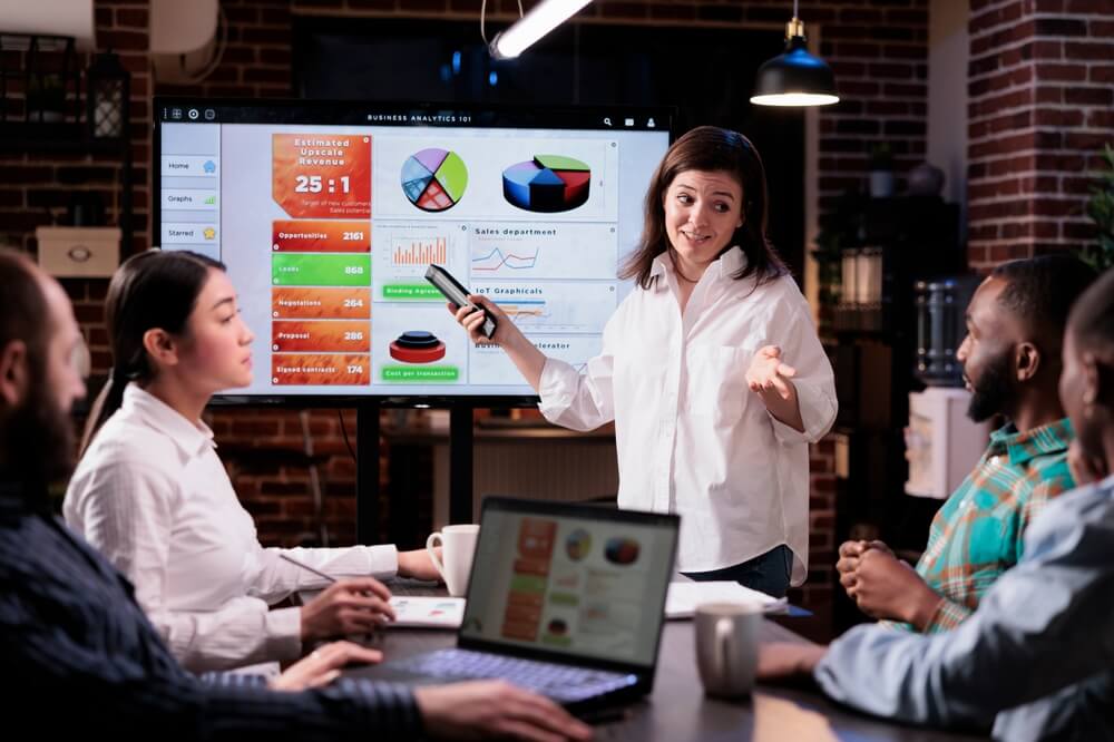 strategic marketing_Smiling caucasian woman in startup office looking at team sitting at desk explaining marketing strategy in late night meeting. Confident employee presenting sales statistics on tv screen.