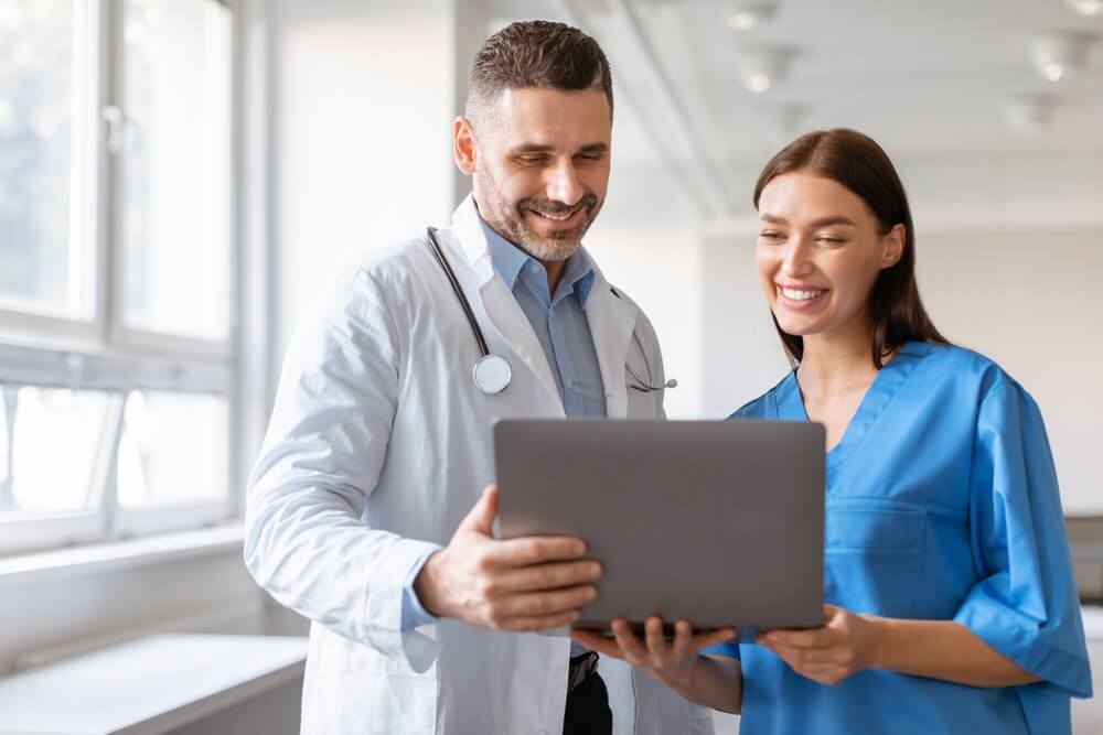 healthcare_Happy male doctor and female nurse talking, working together, using laptop, discussing patient medical checkup results or diagnosis, wearing uniform holding electronic device