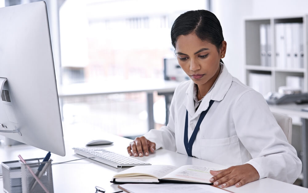 medical seo_Medical research, young doctor with computer and notebook or paperwork sitting at her desk at work. Professional person, results and focused female worker reading paper with pc at her workplace