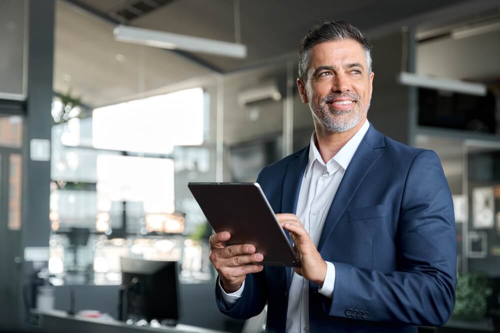 FCMO_Happy middle aged business man ceo wearing suit standing in office using digital tablet. Smiling mature businessman professional executive manager looking away thinking working on tech device.