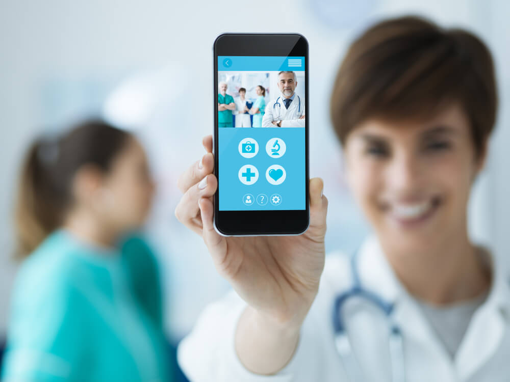 mobile health_Smiling confident female doctor holding a touch screen smartphone and medical staff on the background, medical app concept