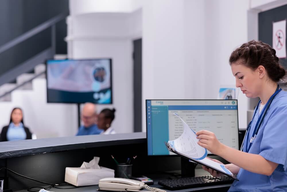 hospital seo_Assistant holding clipboard reviewing the patients medical history before their appointment, working in hospital waiting area. Nurse looking at patient symptoms planning health care treatment