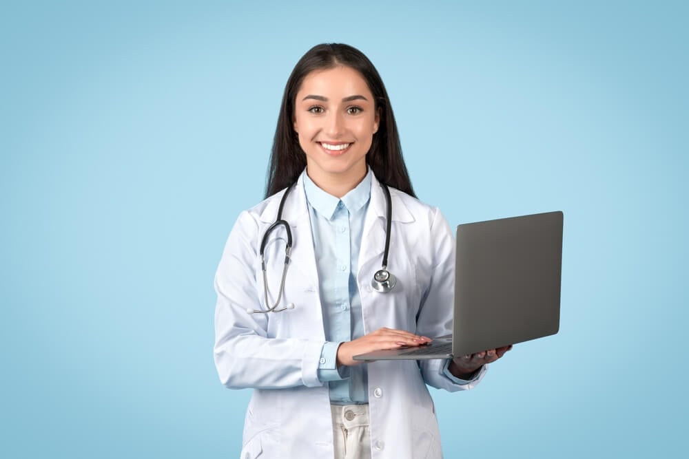 healthcare digital_Professional female doctor holding laptop, representing telemedicine services, with friendly smile and wearing white lab coat, blue background