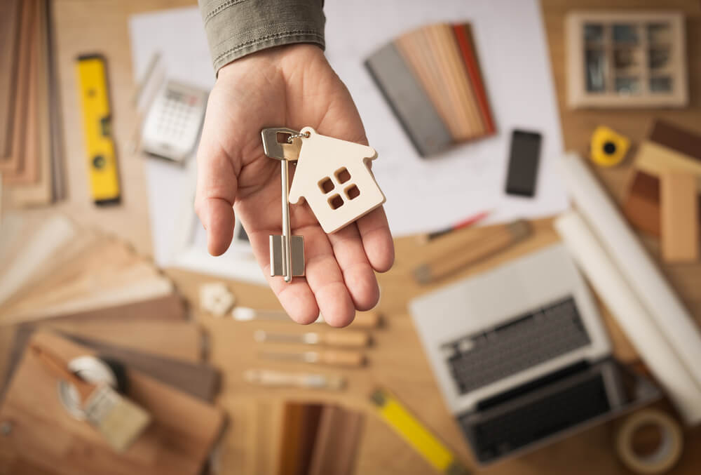 real estate agents_Real estate agent handing over a house key, desktop with tools, wood swatches and computer on background, top view