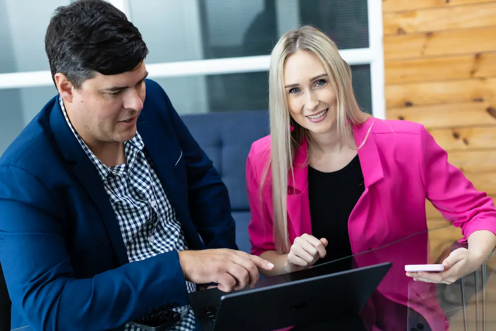 Two colleagues working together at a laptop, man pointing at screen while woman in pink blazer smiles.