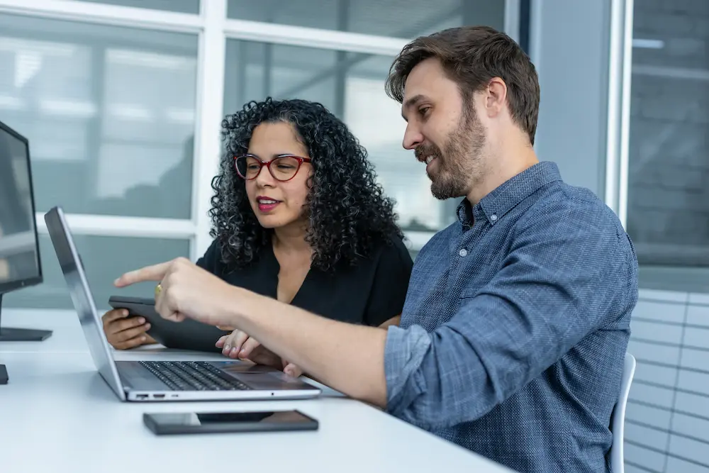 Two colleagues seated at desk collaborating on a laptop, one pointing at screen, the other holding a tablet.